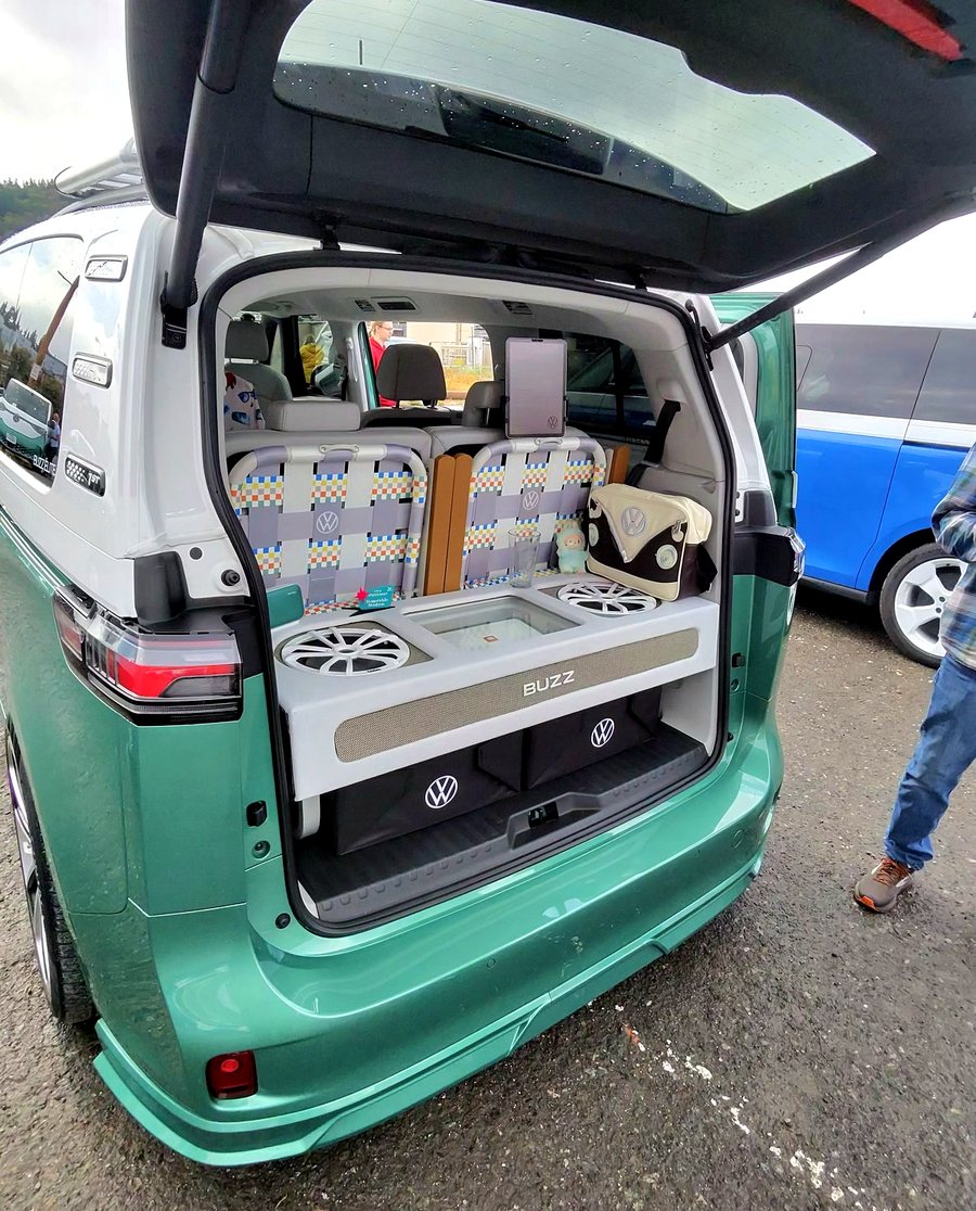 The open tailgate of a lowered teal ID. Buzz showing a custom white cabinet housing two speaker drivers behind wheel-spoke grilles, badged BUZZ across a grille-cloth face, with VW-logo storage bins below.