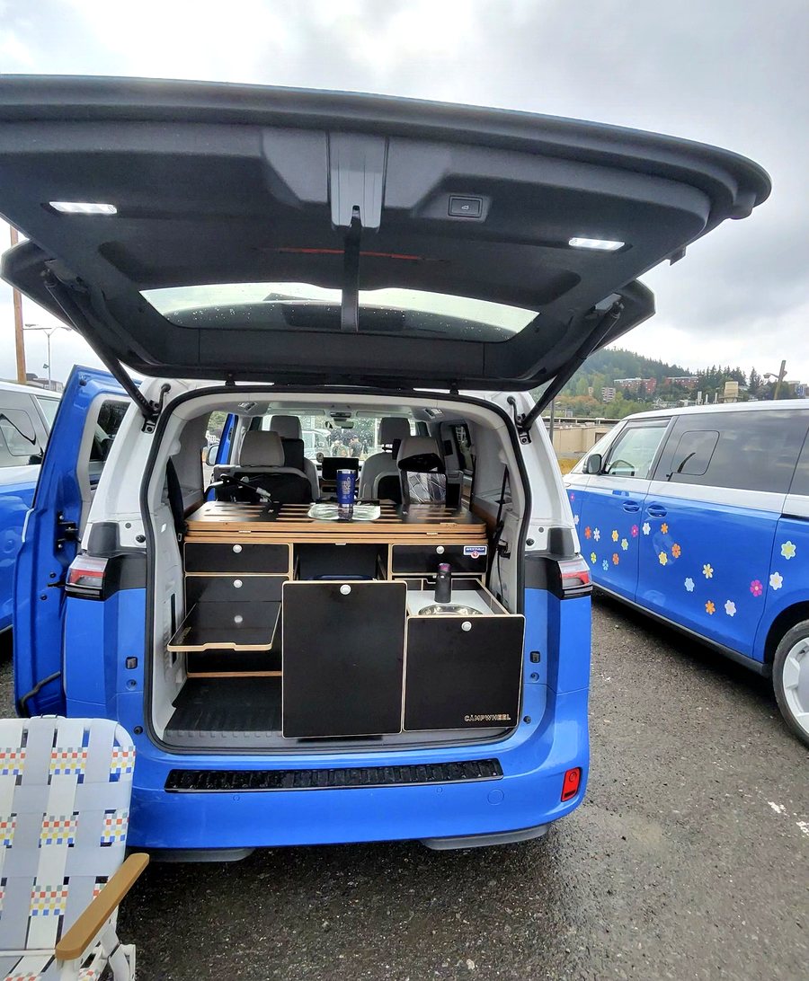 A blue ID. Buzz with the tailgate raised showing a dark cabinet module and birch counter, with a flower-decaled blue Buzz parked alongside.