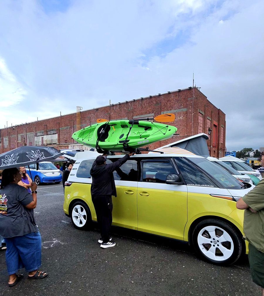 A person lifting a green kayak onto the roof rack of a yellow and white ID. Buzz.