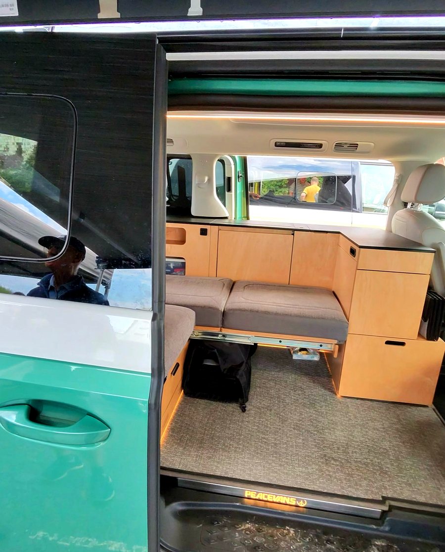 Interior of a green ID. Buzz seen through the sliding door: a grey bench seat facing a full-height birch galley with cabinets, and a Peace Vans sill plate.
