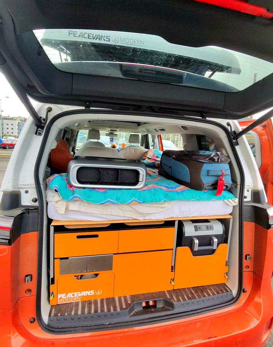 Rear hatch of an orange ID. Buzz open, showing orange cabinet faces, a slide-out fridge and a mattress made up with a patterned blanket.