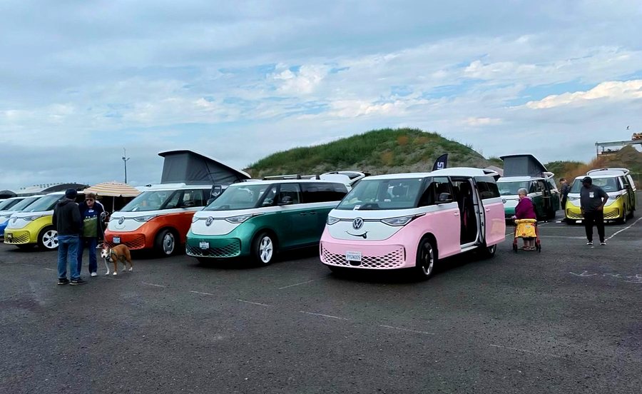 A row of pastel-coloured ID. Buzz vans parked at angles in a gravel lot with a grassy mound behind.