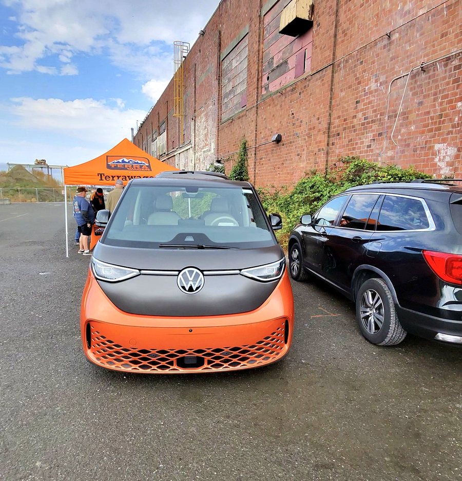 An orange and black ID. Buzz parked in front of a brick warehouse wall, with an orange Terrawagen vendor canopy behind it.